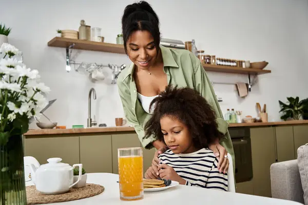 A mother lovingly assists her daughter as they enjoy breakfast together in a bright kitchen. — Stock Photo