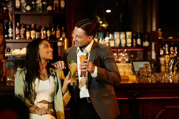 A multicultural couple shares a joyful moment while sipping cocktails at a cozy bar atmosphere. — Stock Photo