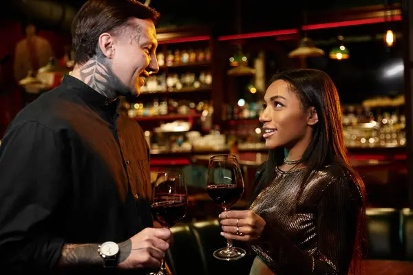Tattooed couple shares a warm connection while toasting with glasses of wine in a busy bar. — Stock Photo