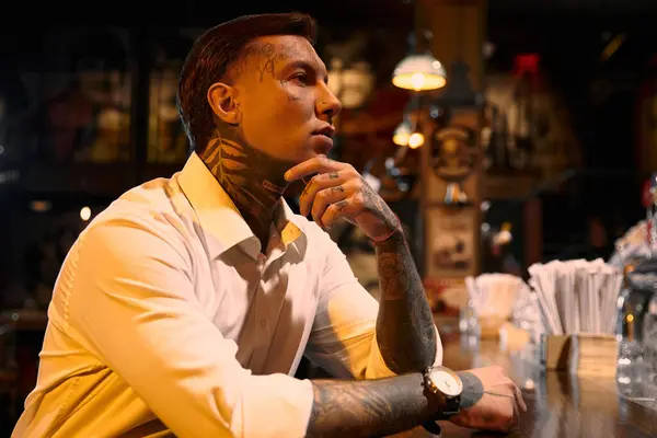A young man with striking tattoos sits thoughtfully at a bar, embracing the evening vibe. — Stock Photo