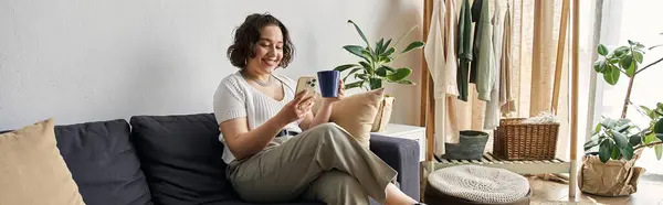 A young woman with curly hair relaxes in her stylish apartment while sipping a drink, banner — Stock Photo