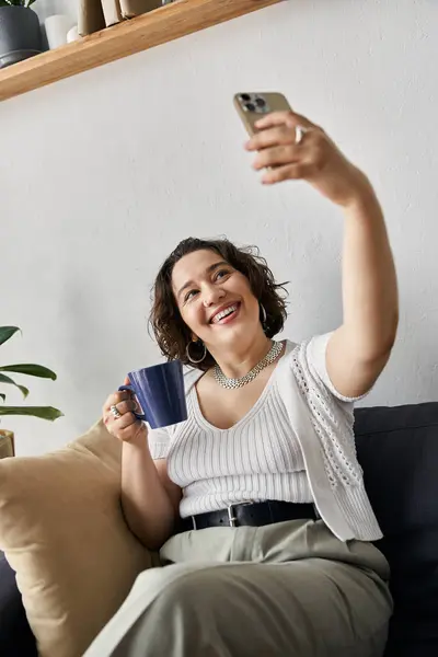 A beautiful young woman enjoys her coffee while taking a selfie in her chic apartment. — Stock Photo