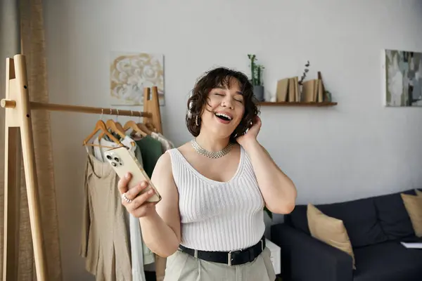 Mujer joven con el pelo rizado sonríe mientras escucha música en su espacio de vida moderno. - foto de stock