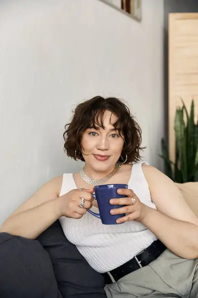 Una bella giovane donna dai capelli ricci si rilassa su un divano, assaporando la sua bevanda in luce naturale. — Foto stock