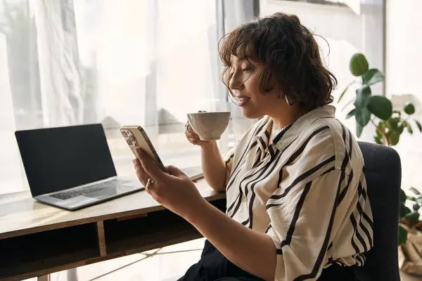 Young woman sips coffee while scrolling on her phone, surrounded by a chic decor. — Stock Photo