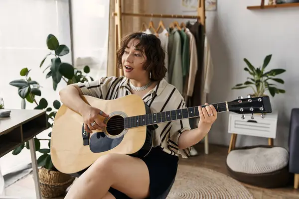 A beautiful young woman with curly hair plays guitar in her modern apartment, exuding joy. — Stock Photo