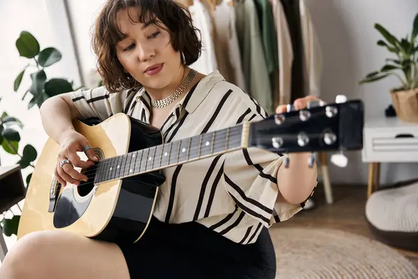 A beautiful young woman with curly hair plays guitar in her stylish apartment, radiating joy. — Stock Photo