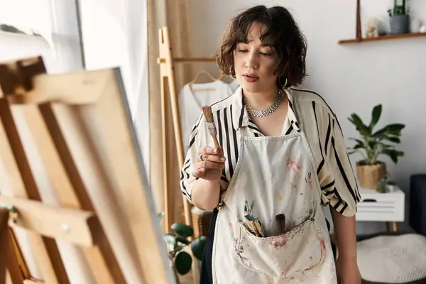 Una giovane donna di talento con i capelli ricci si concentra sulla sua pittura, tenendo un pennello pensieroso. — Foto stock