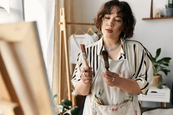 Curly-haired young woman enjoys painting near her easel, surrounded by natural light and plants. — Stock Photo