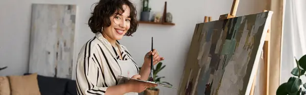 A young woman with curly hair paints on an easel, expressing her creativity in a bright apartment, banner — Stock Photo