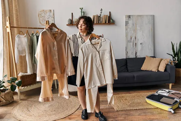 A young woman with curly hair inspects two outfits in her stylish, sunlit apartment. — Stock Photo