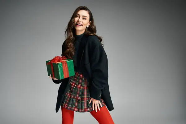 Eccitato giovane donna con i capelli scuri e una gonna a quadri celebra la stagione delle vacanze con un regalo. — Foto stock