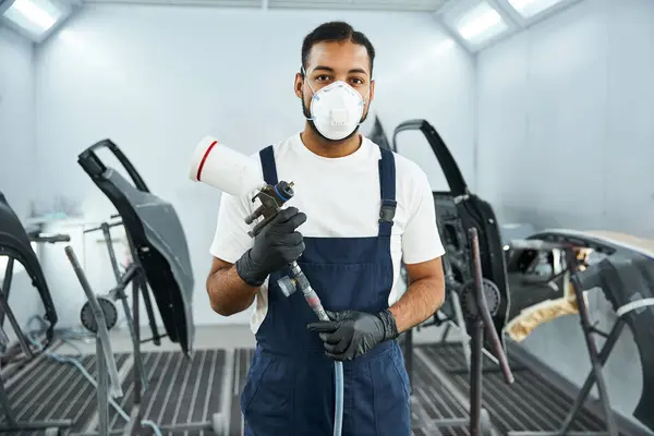 Handsome mechanic wearing a mask and gloves prepares to paint car parts in a workshop. — Stock Photo