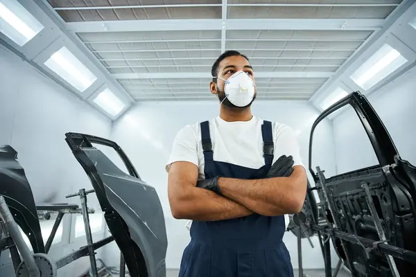 A young mechanic in a mask stands with arms crossed in a busy auto workshop. — Stock Photo