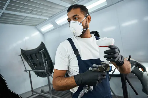 Un joven mecánico en equipo de trabajo prepara una pistola de pulverización para pintar en el taller. - foto de stock