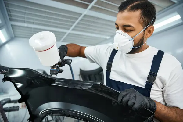 Focused young mechanic spray painting a car part while wearing protective gear and gloves — Stock Photo