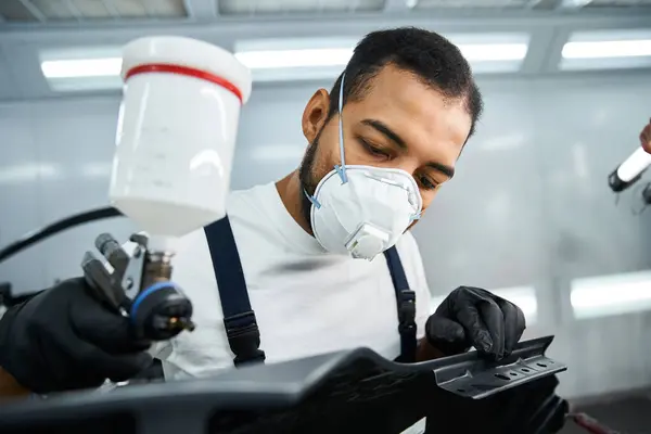 Handsome young mechanic carefully sprays paint on car parts, showcasing expertise in the workshop. — Stock Photo