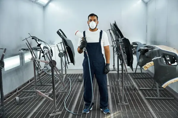 Joven mecánico pinta piezas de automóviles en un taller moderno, vestido con equipo de seguridad. - foto de stock