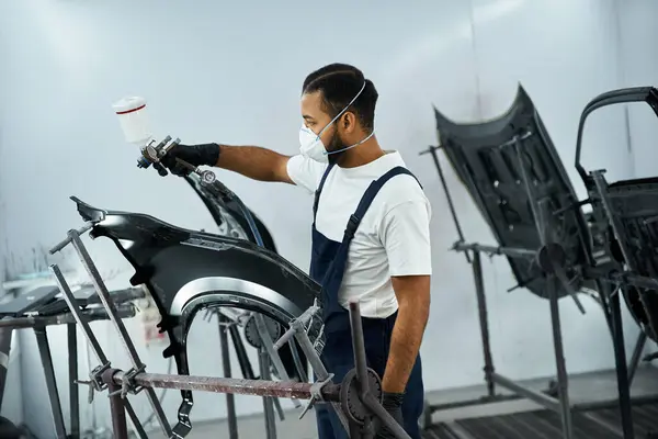 Handsome mechanic expertly applies paint to car parts in a busy workshop during daytime hours. — Stock Photo