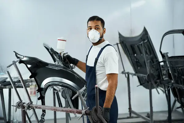 Young mechanic in protective gear paints a car part with focus in a vibrant workshop setting — Stock Photo