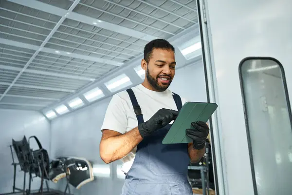 Handsome young mechanic reviews job details on a tablet in the busy workshop during daylight. — Stock Photo