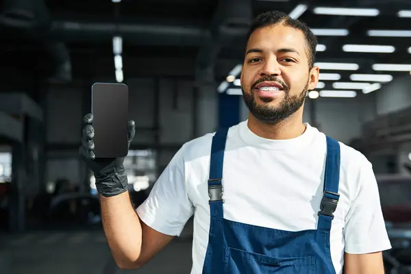 Handsome young mechanic smiles while showcasing a smartphone in a bright workshop setting. — Stock Photo