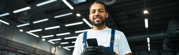 Handsome young mechanic smiles while holding a smartphone in a busy workshop setting. — Stock Photo