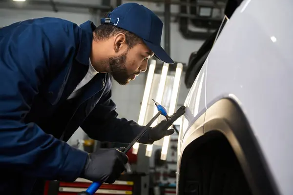 A focused young mechanic repairs a car in a bustling, fluorescent lit workshop. — Stock Photo