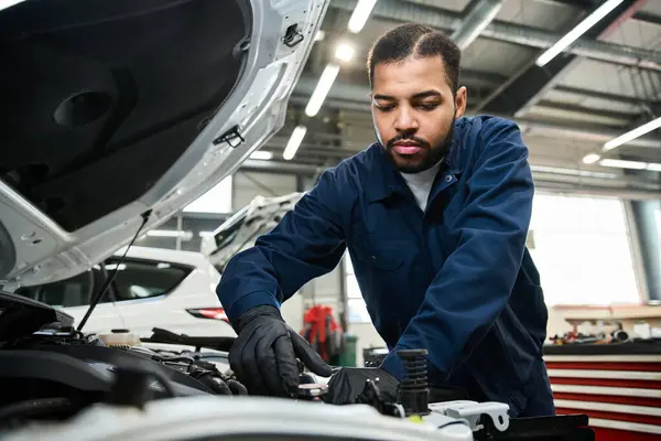 Handsome young mechanic works diligently under the hood of a vehicle in a busy garage. — Stock Photo