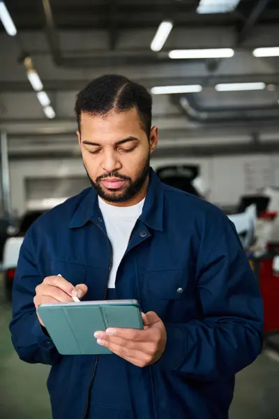 Young mechanic focuses intently on his tablet in a busy workshop, surrounded by vehicles. — Stock Photo