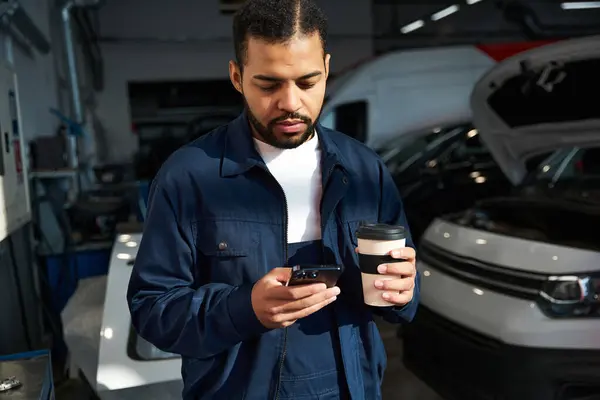 A handsome young mechanic reviews his phone while savoring a warm drink in the garage. — Stock Photo