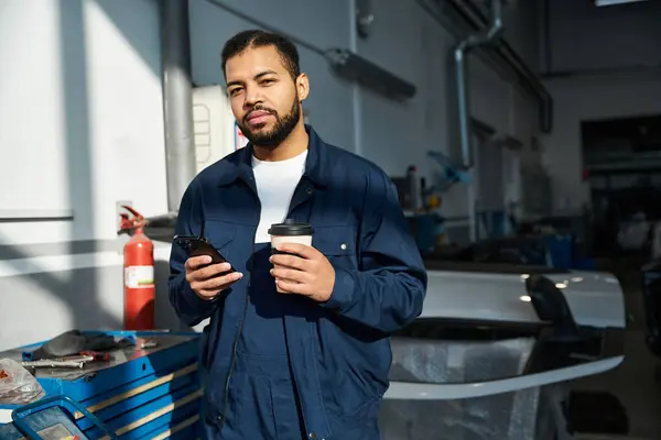 Un bel giovane meccanico si prende un momento per rilassarsi con un caffè e un telefono in garage. — Foto stock