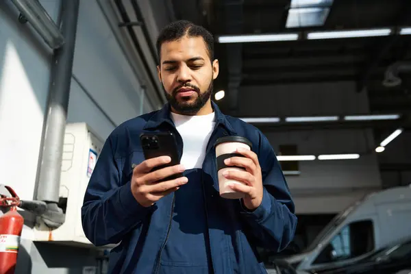 A young mechanic enjoys coffee while engrossed in his phone inside a bustling workshop. — Stock Photo