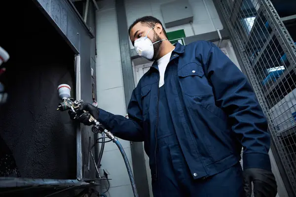 Young mechanic focused on his task while spray painting equipment in a busy workshop. — Stock Photo