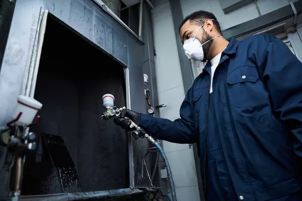 A young mechanic in a blue jacket uses spray equipment with protective gear in a workshop. — Stock Photo