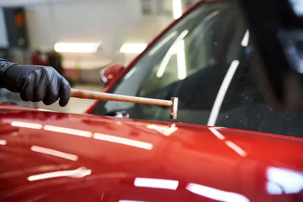 Um mecânico repara um veículo vermelho com um martelo, destacando sua habilidade na garagem movimentada. — Stock Photo