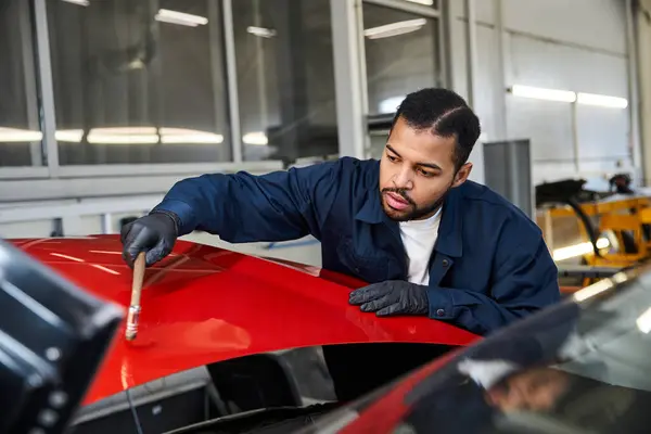 Ein gelernter junger Mechaniker trägt in einer belebten Garage vorsichtig Lack auf ein rotes Auto auf. — Stockfoto