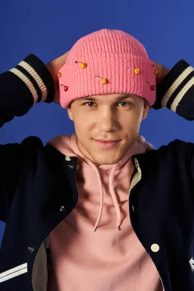 Handsome man in a pink beanie smiles while adjusting his hat in front of a bright blue backdrop. — Stock Photo