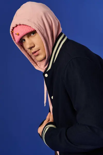 Stylish young man in a pink hoodie poses confidently on blue backdrop. — Stock Photo