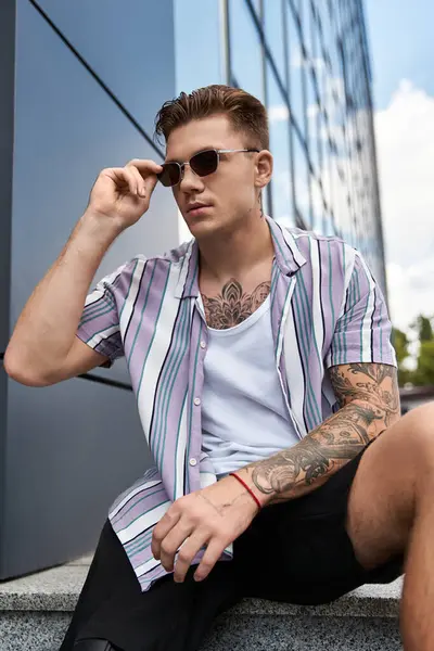 Giovane uomo con tatuaggi si rilassa al di fuori di una struttura contemporanea in una giornata di sole. — Foto stock