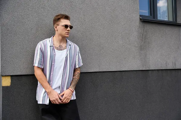 Young man with tattoos stands by a wall, enjoying a sunny day in casual wear. — Stock Photo
