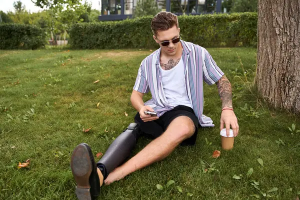 Joven guapo se sienta cómodamente en la hierba, disfrutando de café helado en un entorno tranquilo parque. - foto de stock