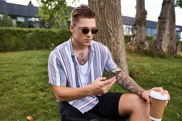 Handsome young man sits on grass, enjoying coffee and using his phone. — Stock Photo