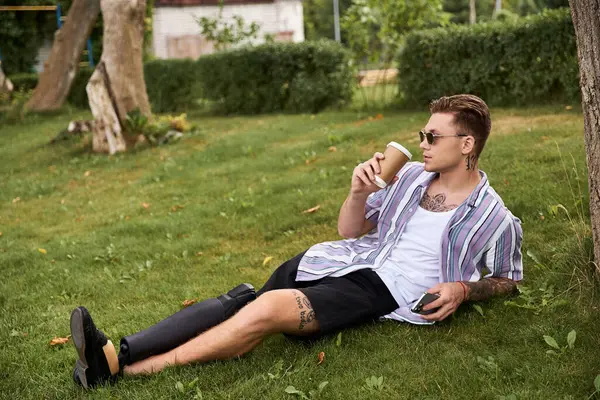 A young man with a prosthetic leg relaxes on the grass, enjoying a drink and sunshine. — Stock Photo
