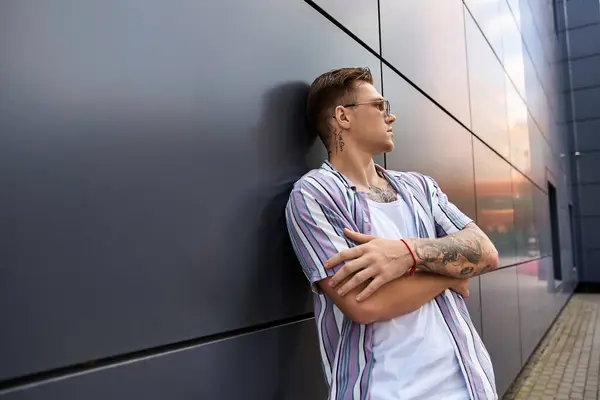 Schöner junger Mann mit Tätowierungen steht selbstbewusst vor einer glatten Wand. — Stockfoto