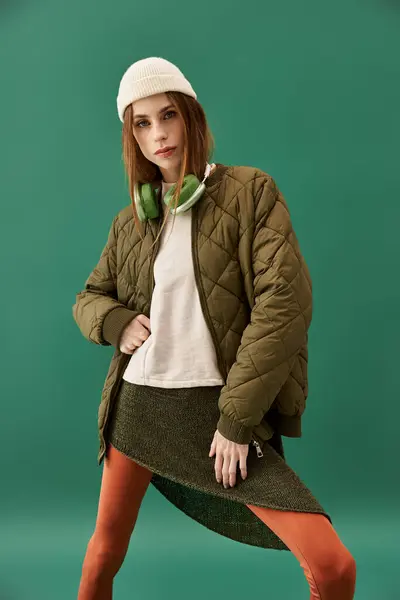 Jeune femme dans une veste vert olive et bonnet pose en toute confiance avec des écouteurs. — Photo de stock
