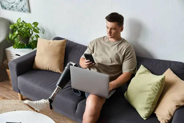 Handsome young man with a prosthetic leg sitting on a sofa, focused on his laptop and smartphone. — Stock Photo