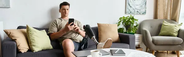 Young handsome man sits comfortably on a sofa, engaged with his smartphone in a bright room. — Stock Photo