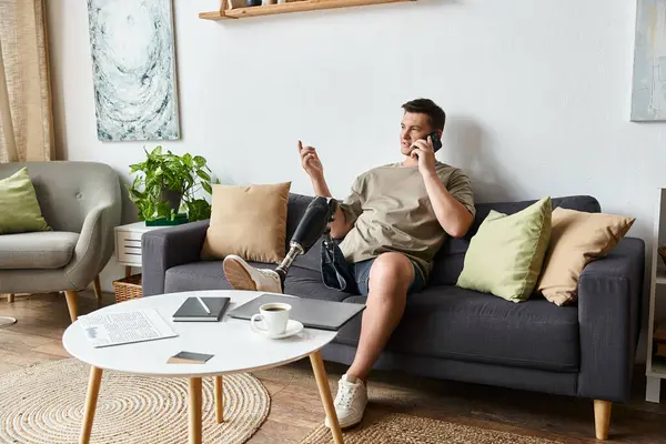 Giovane uomo con una gamba protesica gode di una telefonata mentre si rilassa su un accogliente divano all'interno. — Foto stock