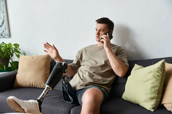 Joven hombre guapo con una pierna protésica se sienta en un sofá mientras habla en su teléfono inteligente. - foto de stock
